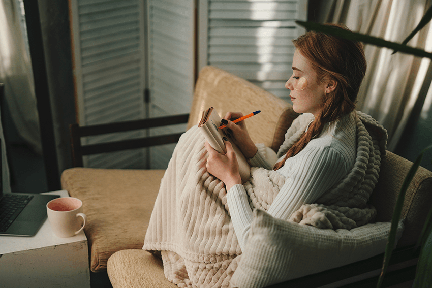 Extract Labs Holiday Gift Guide 2023 8 image of a woman sitting on a couch journaling bundled up in a blanket for blog about Extract Labs Holiday Gift Guide 2023 | CBD Gifts | Pet Gifts | Dog Gifts | Cat Gifts | Gifts for Cat lovers | Gift guide 2023