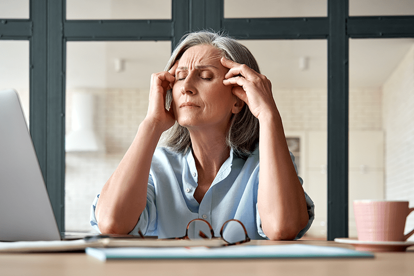 image of an older woman rubbing her head in front of her computer for the blog about CBD for Menopause Symptoms & 3 Cannabinoids that May Help