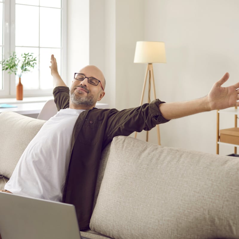Happy adult man who has finished his work is sitting on sofa at home with notebook and laptop computer, smiling with eyes closed and feeling relaxed and satisfied