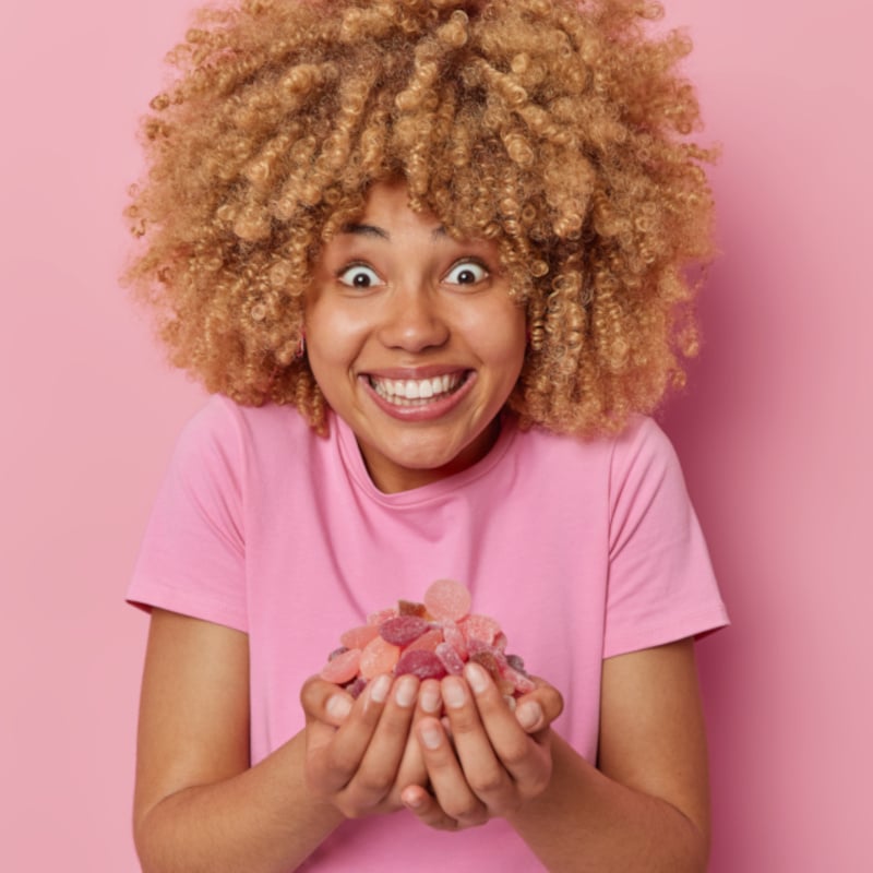 Happy woman holds delicious sweet jellys in hands smiles toothily glad to eat favorite dessert dressed in casual t shirt isolated over pink background. Cheerful girl eats yummy sugary snack.