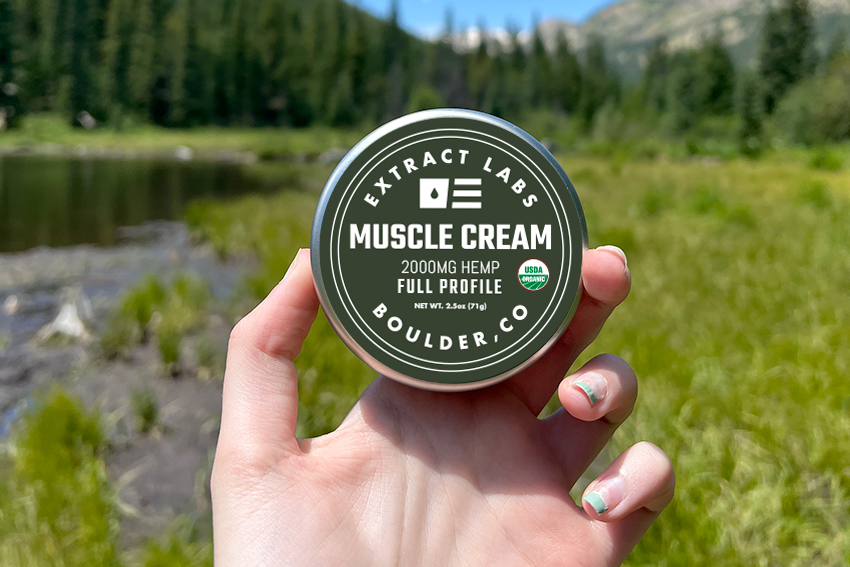 image of Organic Hemp Muscle Cream being held in front of a mountain scenery