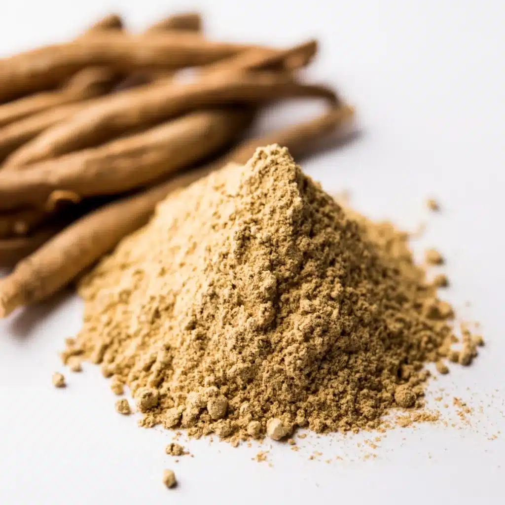 Pile of tan ashwagandha root powder beside dried ashwagandha roots on a white surface
