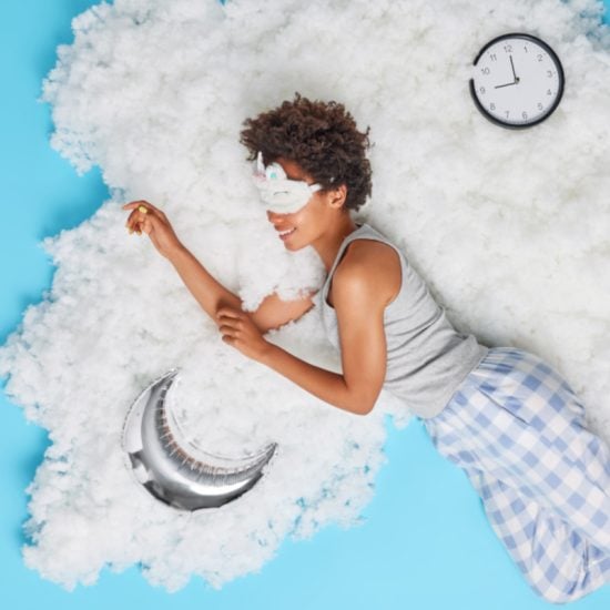 Positive relaxed woman takes nap during daytime poses bare feet in pajama smiles happily wears sleepmask on eyes poses on white cloud with inflated moon and clock showing time. Blue background